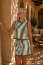 A woman stands in a sunlit courtyard wearing a sleeveless olive green and white striped dress with a matching mini skirt. The setting features textured stucco walls, arches, and outdoor seating in the background.