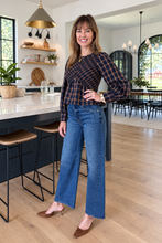 A woman stands in a bright, modern kitchen wearing a dark brown and navy plaid long-sleeve top with blue wide-leg jeans and brown pointed-toe heels.