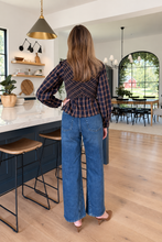 A woman stands with her back to the camera in a bright, modern kitchen, wearing a dark brown and navy plaid wrap-style long-sleeve top and wide-leg blue jeans.