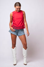 A woman modeling a sleeveless bright red top with fringe trim, paired with denim shorts and white cowboy boots, against a light gray studio background.