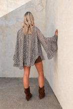 A woman stands with her back to the camera wearing a loose zebra-print cardigan, brown shorts, and brown ankle boots, posing in a corner with textured beige walls.
